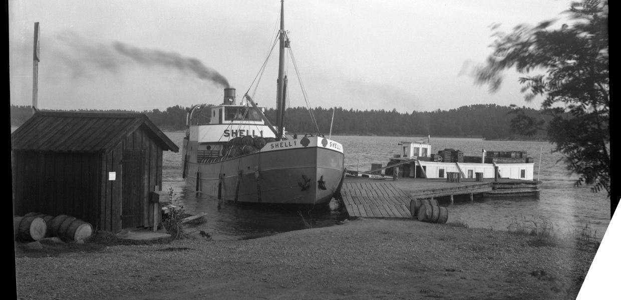 A large boat with the name 'Shell 1' on the side lies in water beside a dock and wooden shed