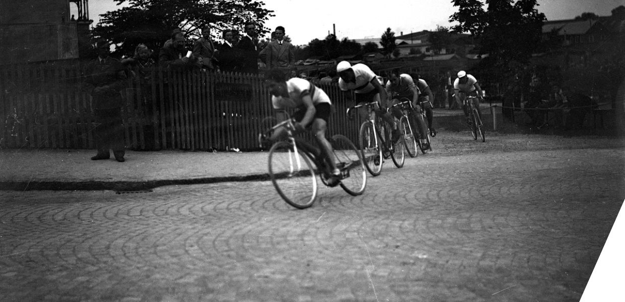 Men taking part in a cycling race moving around a corner on a paved street. People look on from behind a fence. 