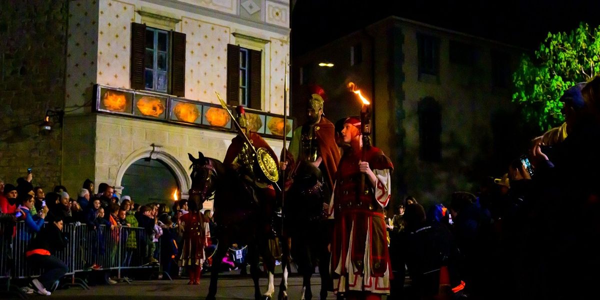 Night parade with people in Roman costumes, one on horseback, holding torches, watched by a crowd behind barriers.