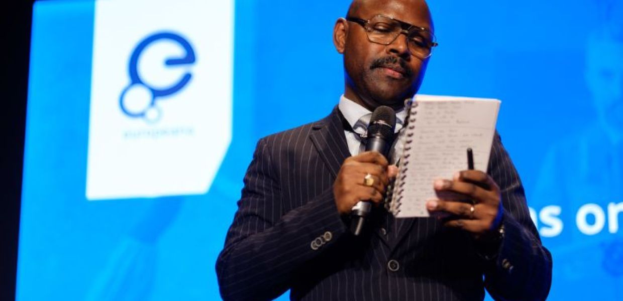 Mr Gee holding a microphone looking at a notebook, with the Europeana logo in the background.