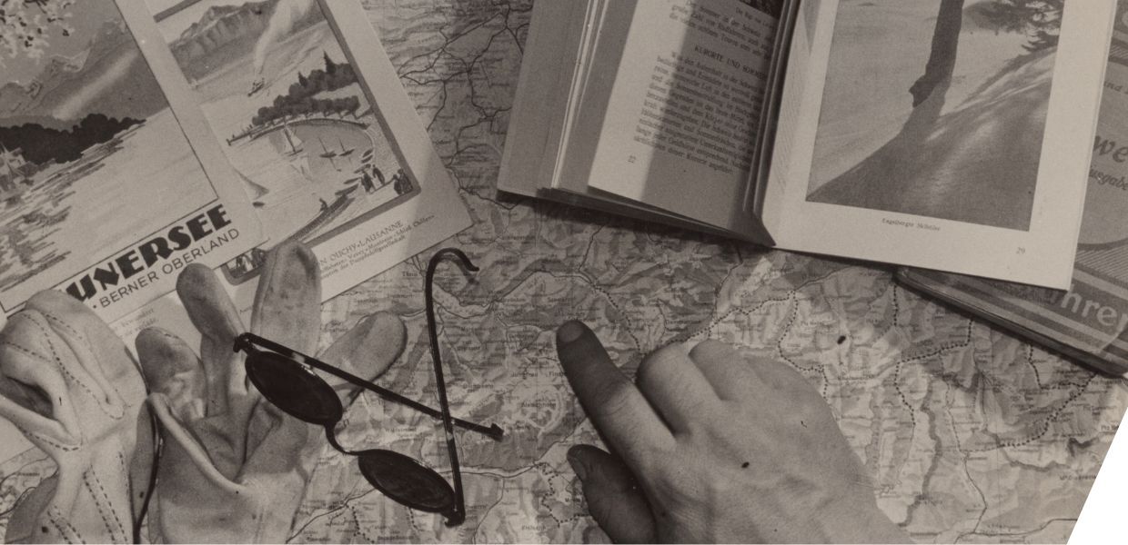 This image shows a travel planning scene for the Swiss Alps, specifically the Berner Oberland region. It includes a map, guidebook, postcards, gloves, sunglasses, and a hand pointing to a spot—likely around Thun or Interlaken.