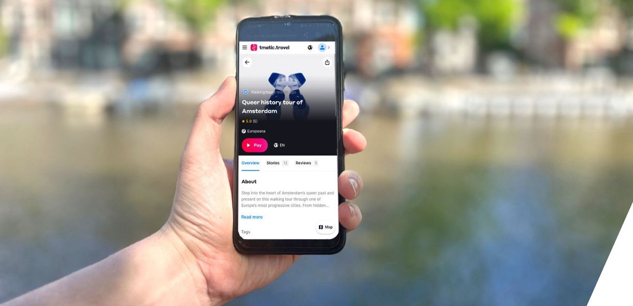 A hand holding a smartphone displaying a queer history walking tour of Amsterdam, with a scenic canal and blurred buildings in the background.