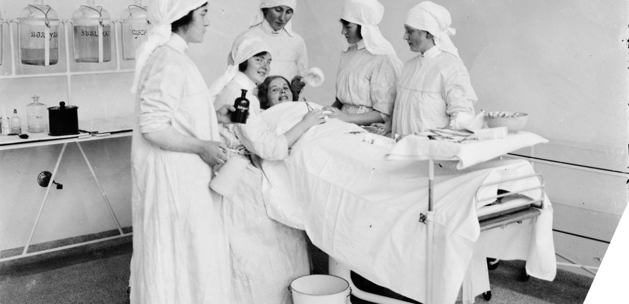 A group of nurses and a patient in a historical hospital setting, with the patient lying on a surgical bed and the nurses gathered around her, smiling.