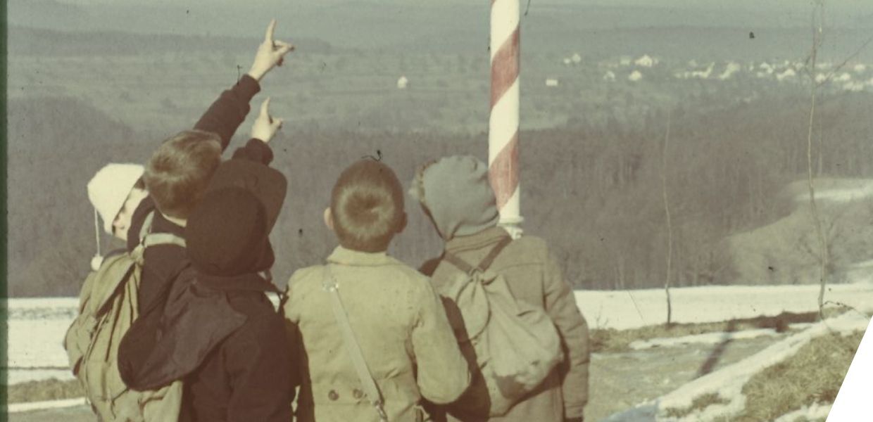 Four children wearing backpacks point toward a tall striped pole in a snowy, rural landscape.