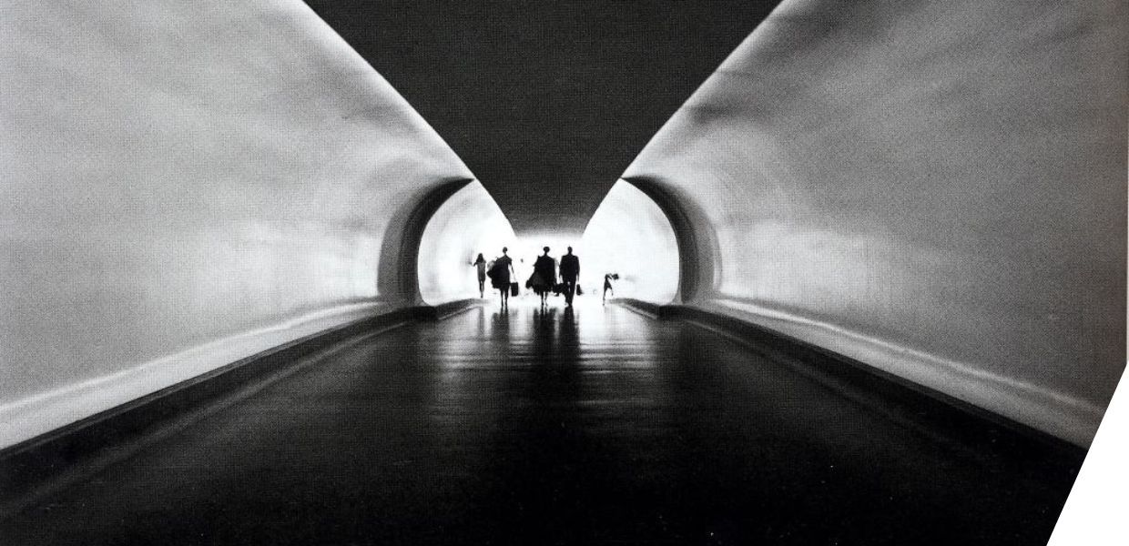 This black-and-white image features a dramatically lit tunnel, with smooth, curved walls that converge towards a bright light at the far end. The architectural perspective draws the viewer’s eye to the center, where silhouettes of several people and a dog are walking toward the light.